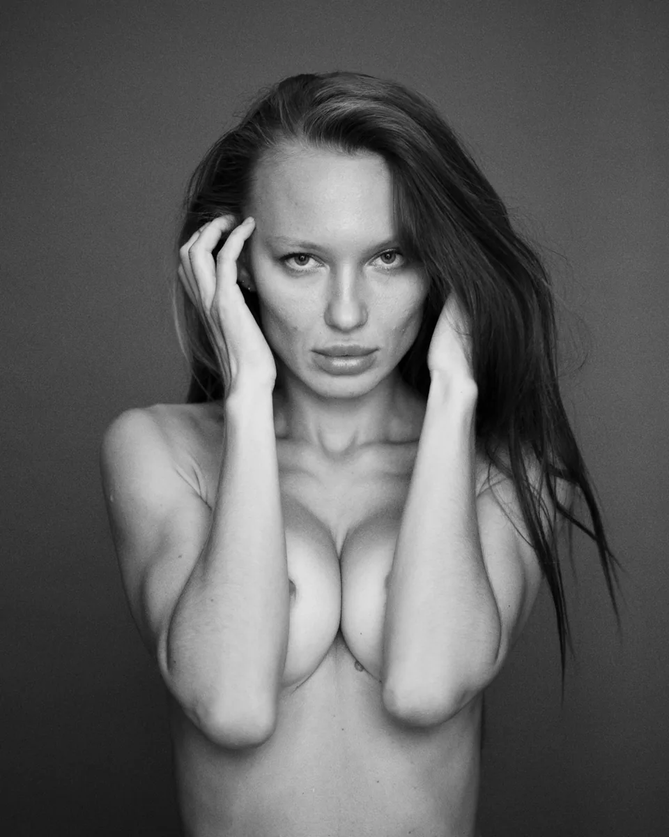 A black-and-white nude portrait of a woman with her hands on her head