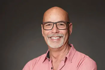 Laughing man with glasses, a white beard and a pink shirt against a dark background