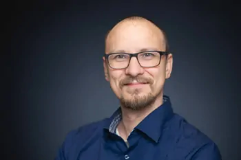 Man with glasses, a short beard and a blue shirt against a dark background