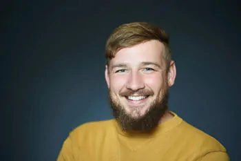 Smiling bearded man in a mustard-yellow sweater against a dark grey background