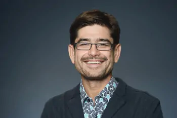 Smiling man with dark hair, glasses and a patterned shirt under a dark jacket