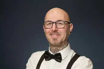 Man with glasses, a bow tie, suspenders and a visible neck tattoo against a dark background