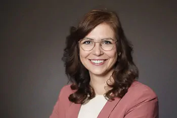 Smiling woman with glasses, brown curls and a pink blazer against a dark background